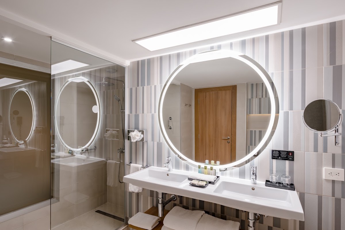 Modern bathroom with dual illuminated round mirrors, white countertop with twin sinks, striped tile walls, and walk-in shower at Hotel Riu Palace Antillas.