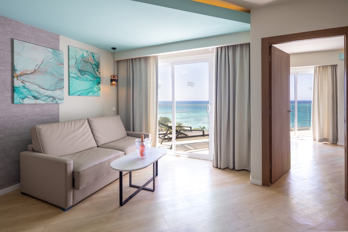 Beachfront suite at Hotel Riu Palace Antillas with turquoise ocean view through glass doors, gray sofa, and modern decor.