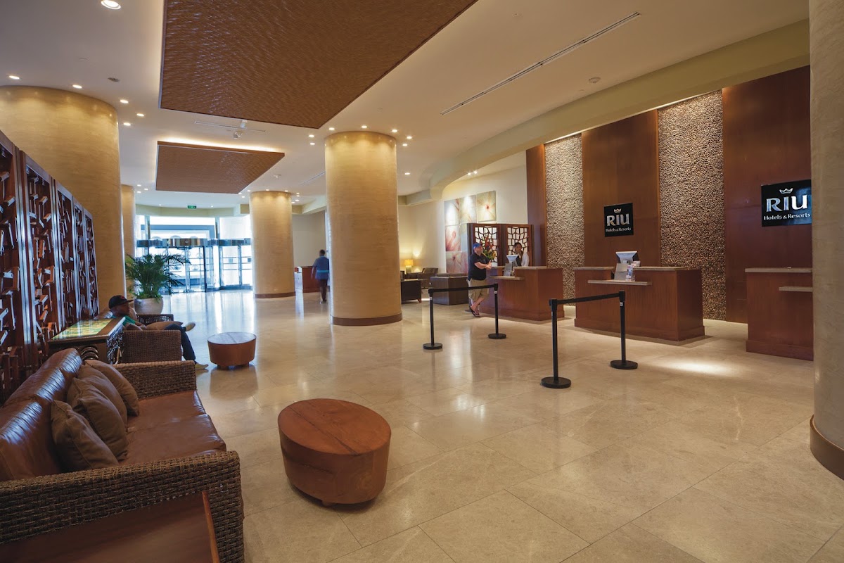 Spacious hotel lobby at Riu Palace Antillas with beige tile flooring, brown wooden columns, and reception desks along textured walls.