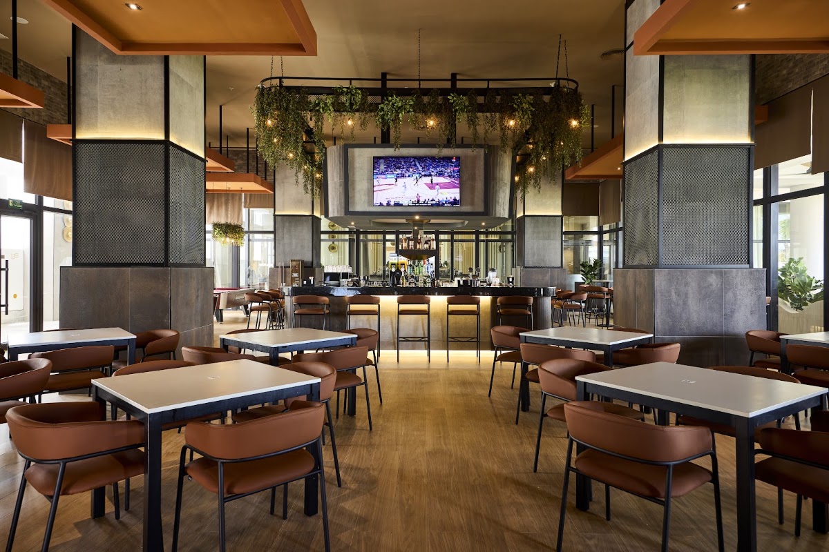 Modern dining area with tan tables, brown leather chairs, and a central bar with hanging greenery and TV screen above.