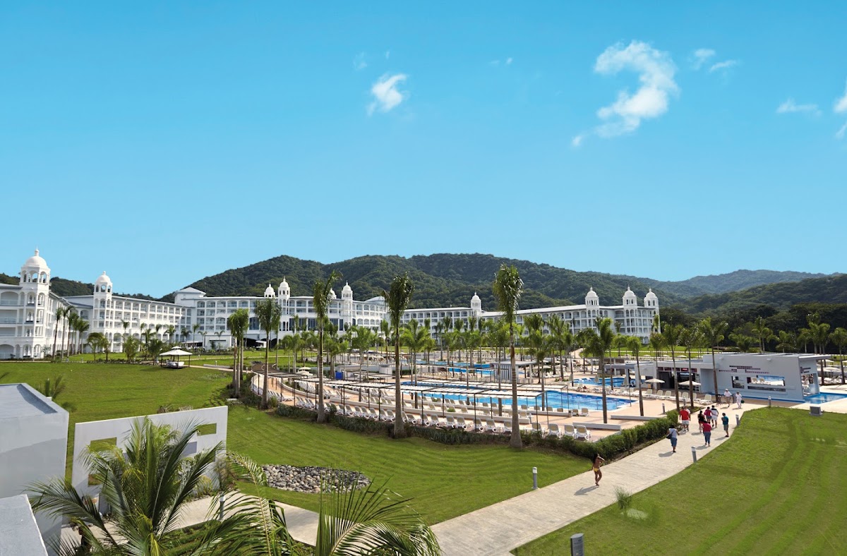 White resort buildings with domed towers overlook manicured lawns, multiple pools with palm trees, and forested mountains under a clear blue sky.