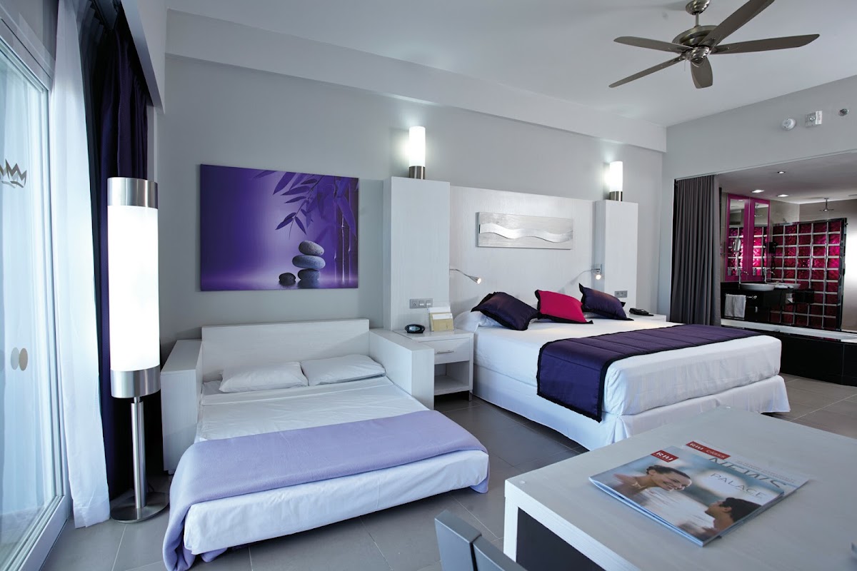 Modern resort room with two beds, purple artwork, white furniture, ceiling fan, and red brick accent wall visible through glass doors.