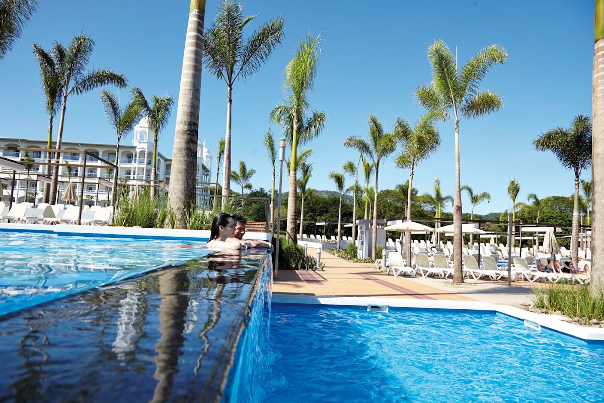 Couple relaxing in a swimming pool at Hotel Riu Palace Costa Rica, with palm trees, lounge chairs, and white resort buildings in the background.