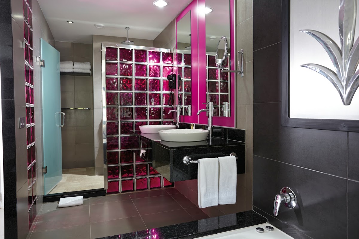 Modern bathroom with dual white vessel sinks, magenta glass tile accent wall, black countertop, and walk-in shower at Hotel Riu Palace Costa Rica.