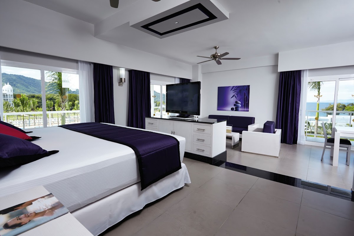 Modern resort bedroom with king bed, flat-screen TV, and seating area overlooking ocean and forested mountains through floor-to-ceiling windows.