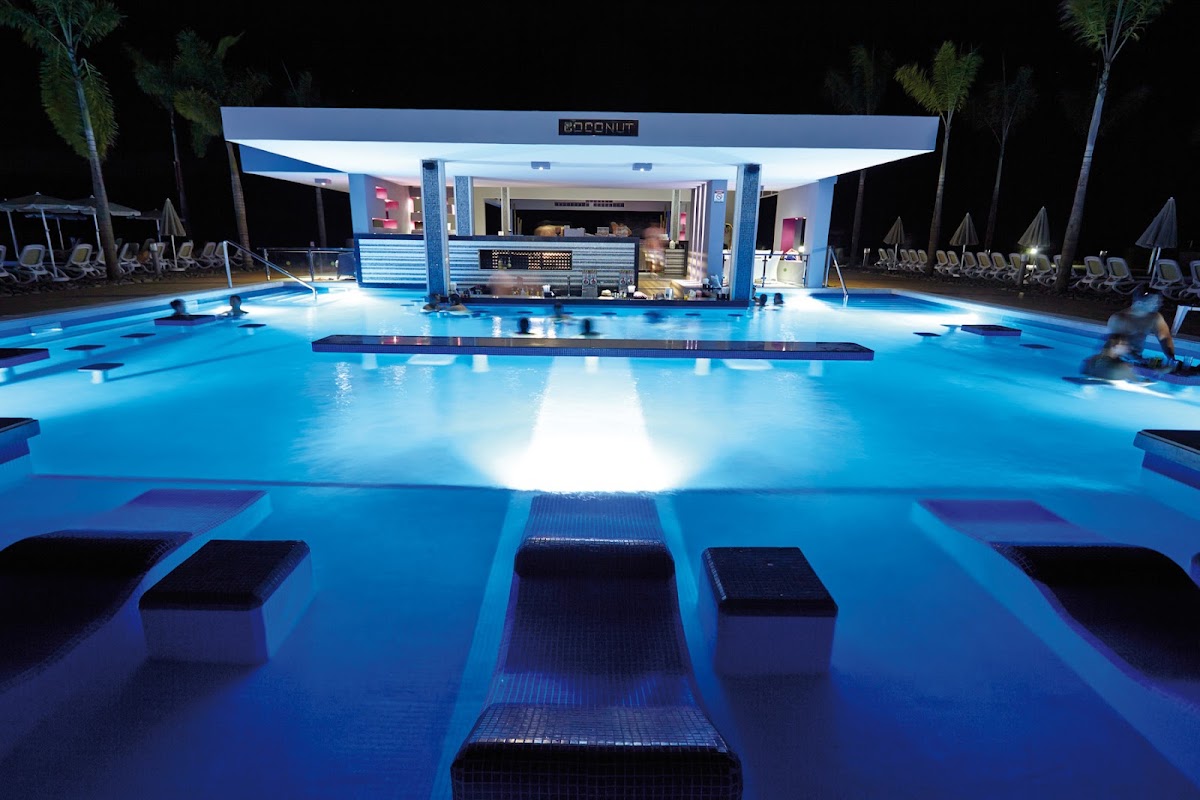 Nighttime view of an illuminated pool at Hotel Riu Palace Costa Rica with a lit bar structure, loungers, and palm trees surrounding the water.