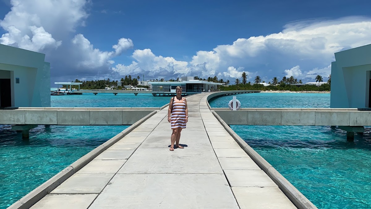 A guest walks along a concrete pier over turquoise lagoon water toward the Hotel Riu Palace Maldivas, with palm trees and resort buildings visible across the water.