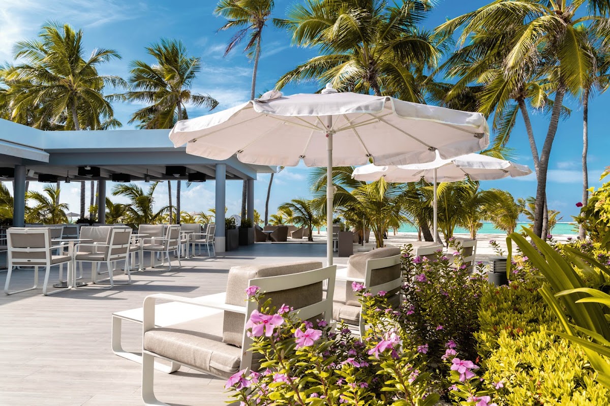 Beach deck at Hotel Riu Palace Maldivas with white umbrellas, lounge chairs, pink flowers, palm trees, and turquoise ocean beyond.