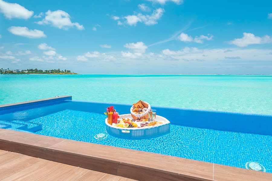 Floating breakfast platform with tropical fruits and drinks in a resort pool overlooking turquoise lagoon and palm-lined island.