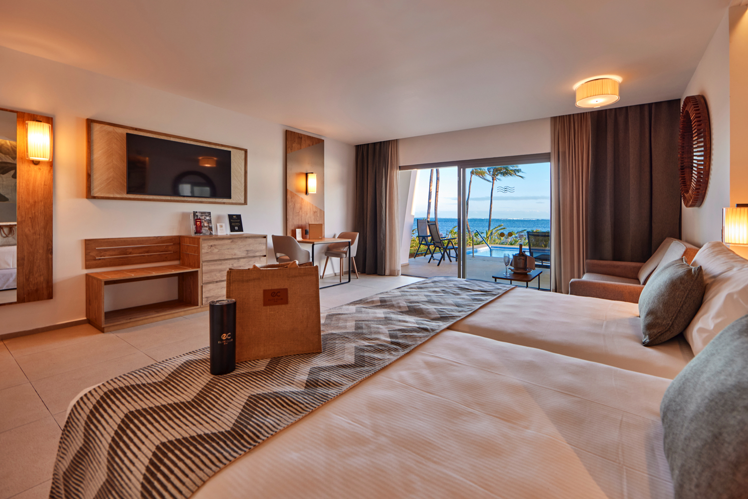 Spacious resort room with two beds, wood furnishings, ocean view from balcony, and pool visible beyond palm trees.