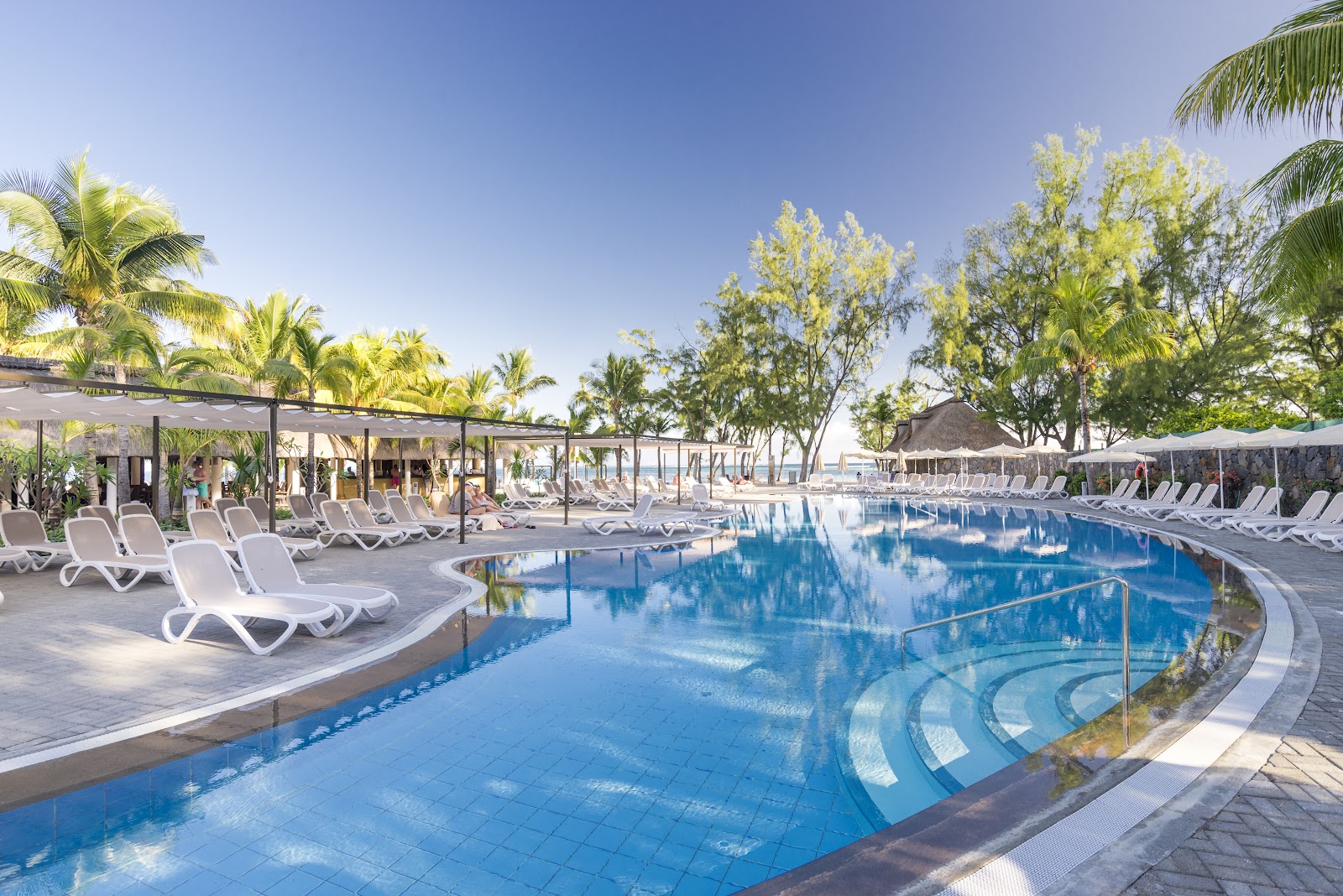 Resort pool with curved design, white loungers, black shade structures, palm trees, and ocean view under clear sky.