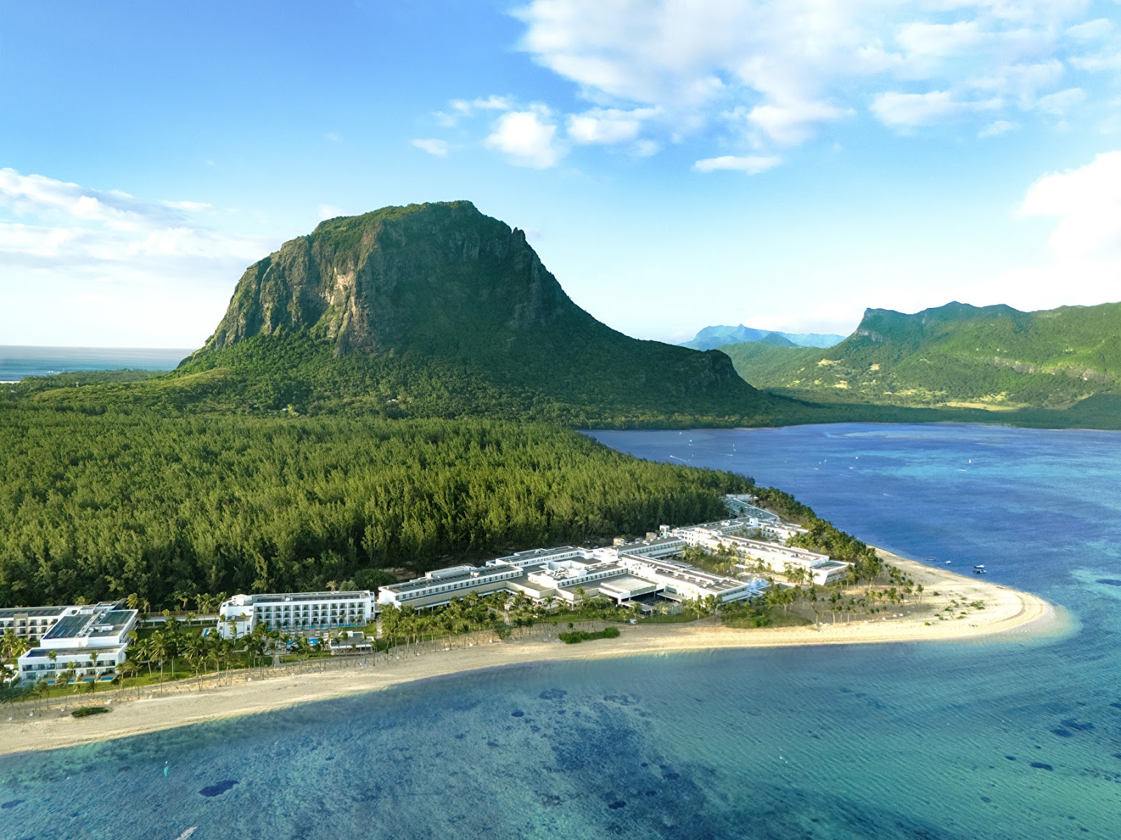 Resort buildings sit on a sandy beach beside turquoise lagoon waters, with dramatic volcanic peaks and lush green mountains rising behind.