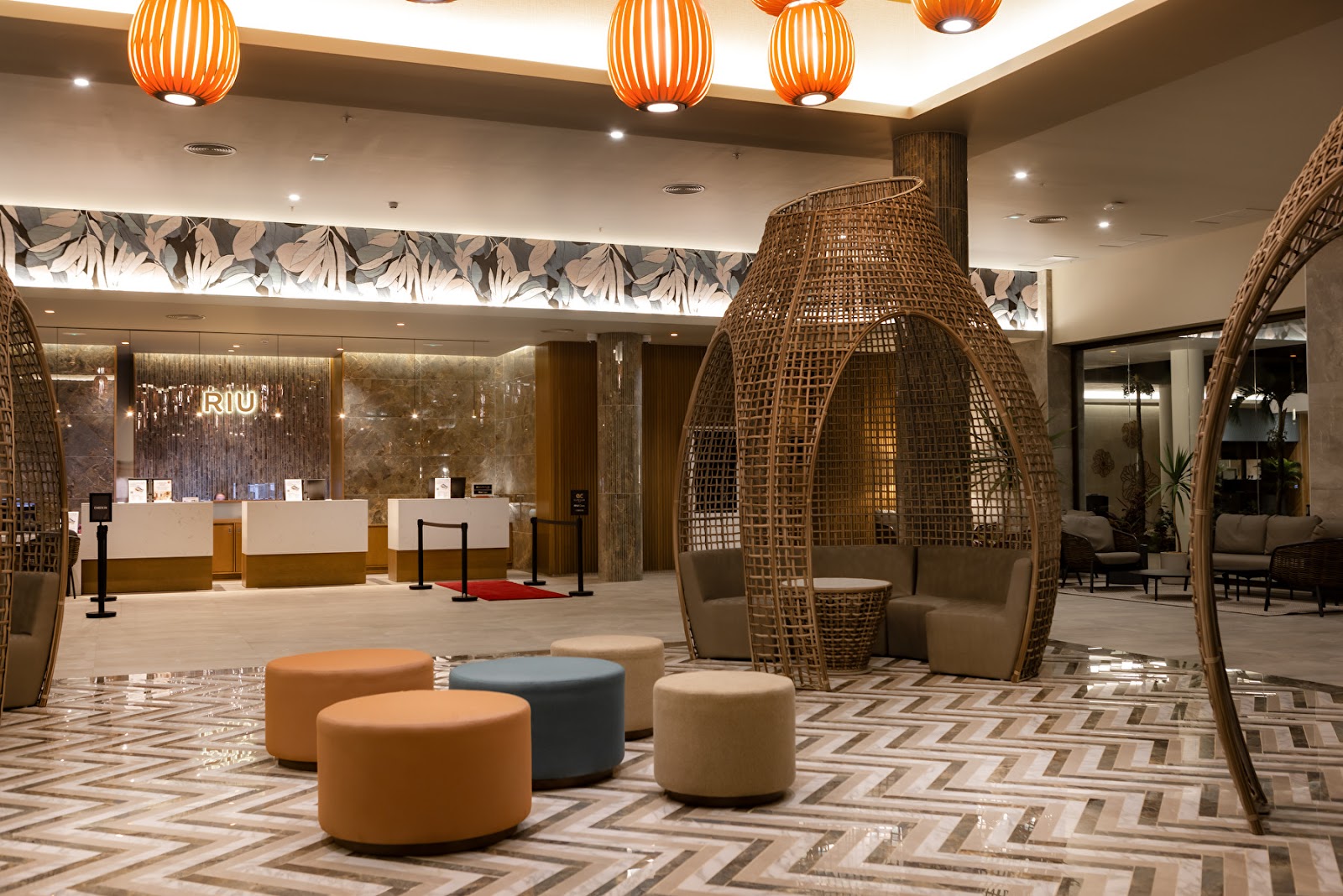 RIU Palace Mauritius lobby with woven pod seating, orange pendant lights, and check-in desks beneath tropical leaf artwork.
