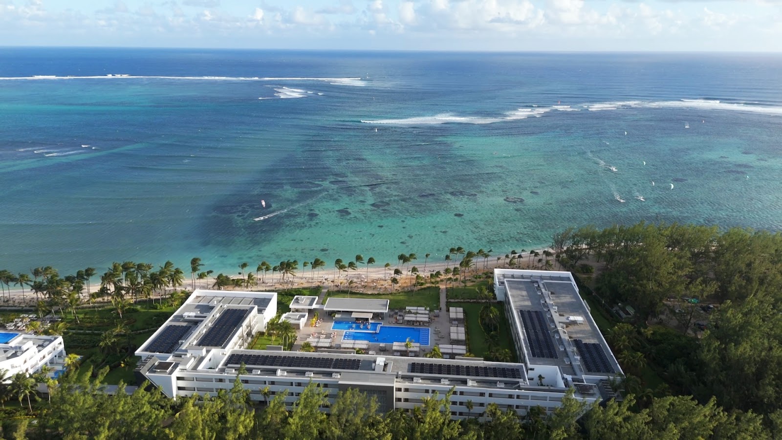 Resort pool with lounge chairs and palm trees, white building structures behind, mountains visible under blue sky with clouds.