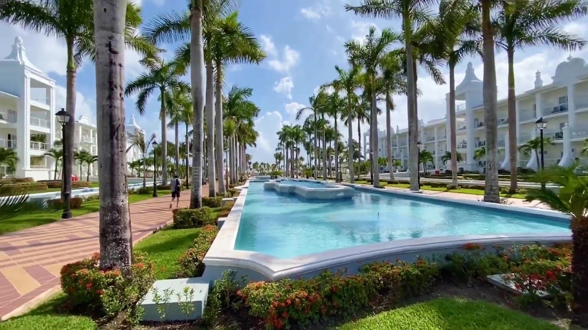 Palm-lined resort grounds with a rectangular pool surrounded by manicured lawns and white colonial-style buildings under blue sky.