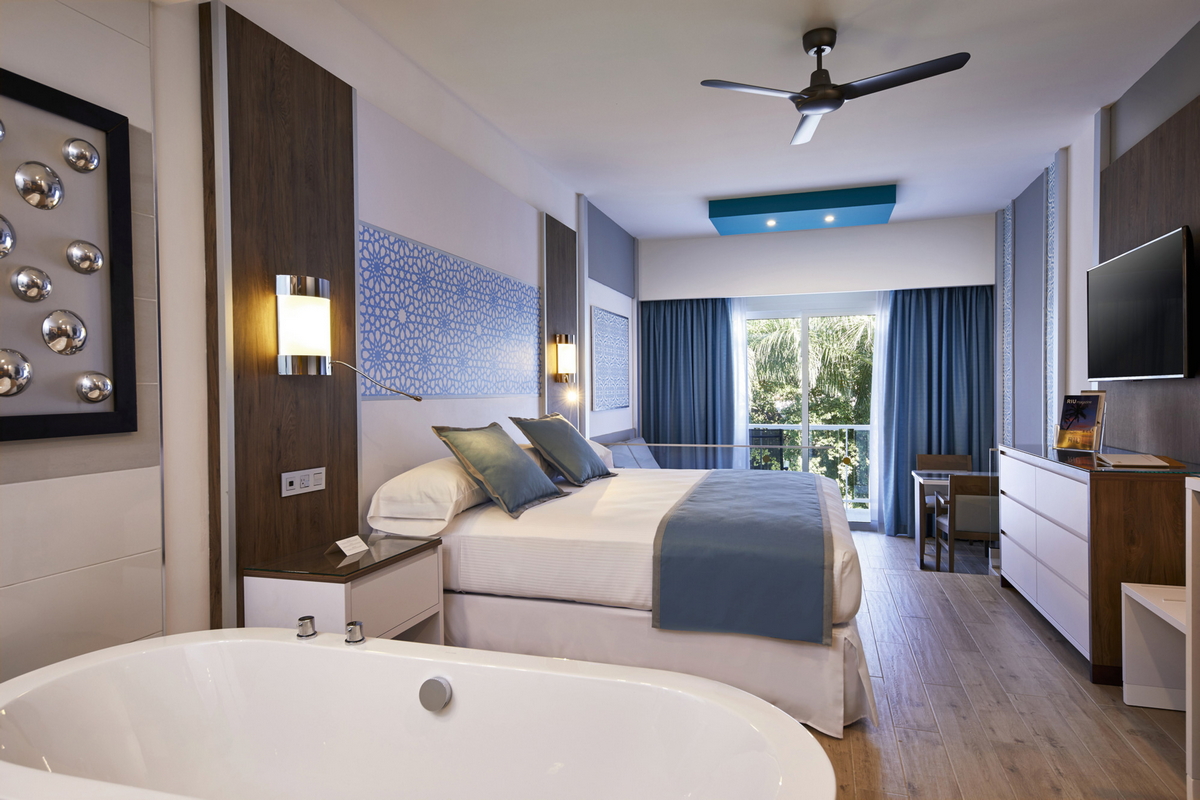 Modern resort bedroom with soaking tub in foreground, queen bed with blue accents, dark wood furnishings, and balcony overlooking tropical grounds.