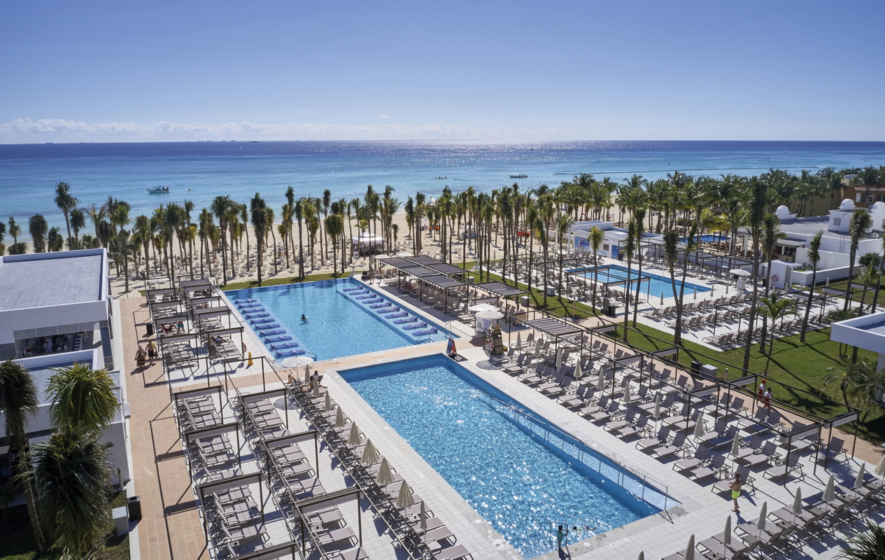 Multiple swimming pools with loungers and palm trees facing the turquoise Caribbean Sea at Hotel Riu Palace Riviera Maya.