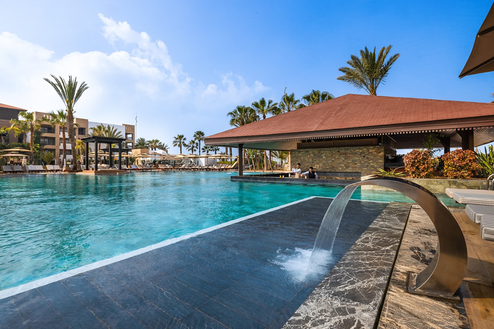 Large turquoise pool with waterfall feature and modern stone pavilion, palm trees and hotel buildings visible under clear blue sky.