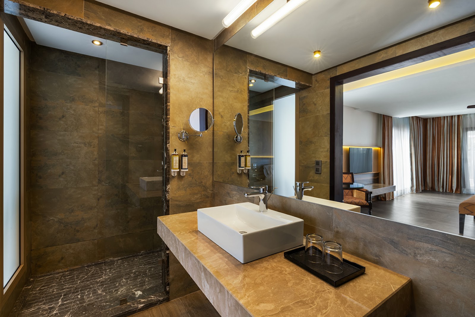 Luxury bathroom with golden marble walls, white rectangular sink, walk-in shower, and view into bedroom at Hotel Riu Palace Tikida Agadir.