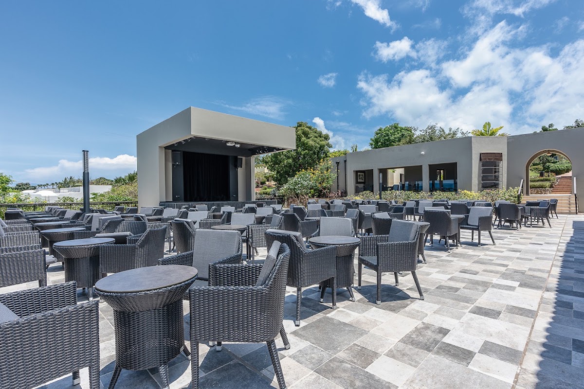 Outdoor terrace with gray wicker seating and round tables facing a modern stage building under blue sky at Hotel Riu Palace Zanzibar.