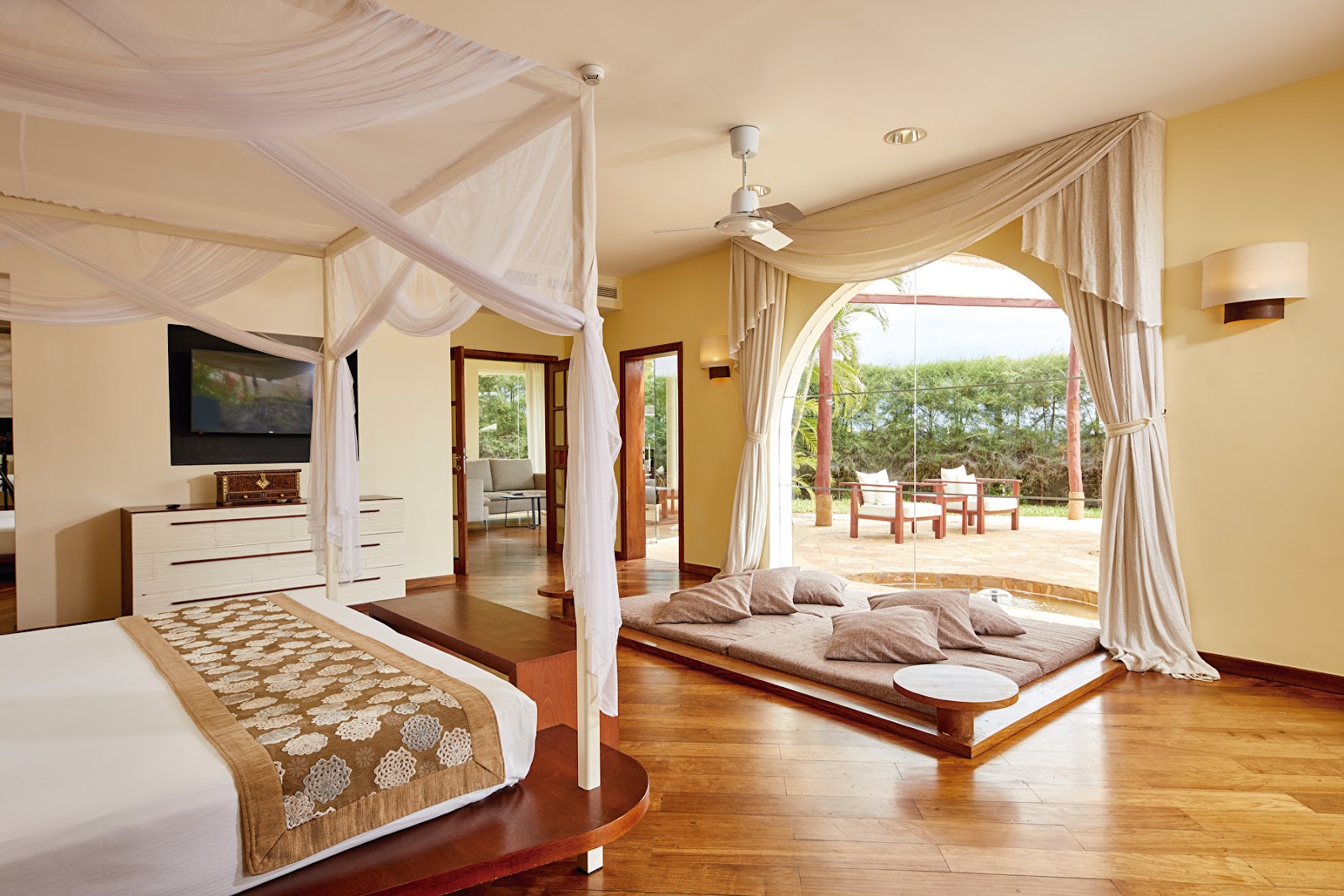 Spacious suite at Hotel Riu Palace Zanzibar with two canopy beds, yellow walls, wooden floors, and arched doorway opening to terrace with lounge chairs.