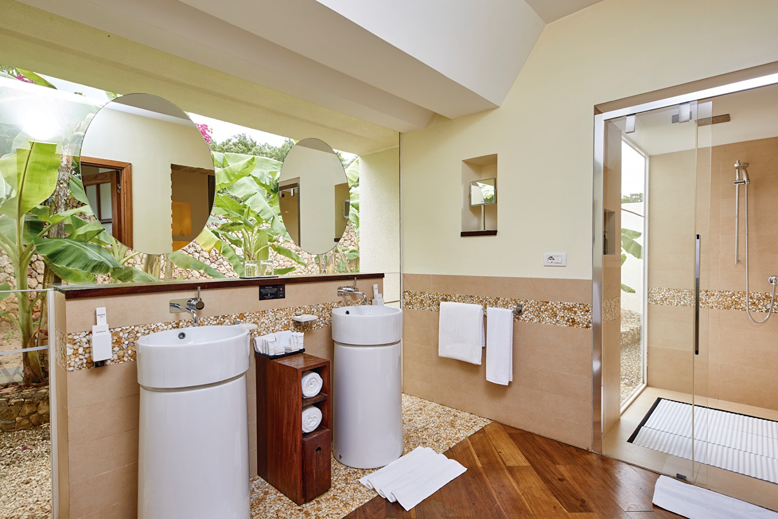 Spacious bathroom with dual cylindrical sinks, large mirror, pebble tile accents, walk-in shower, and tropical plants visible through openings at Hotel Riu Palace Zanzibar.