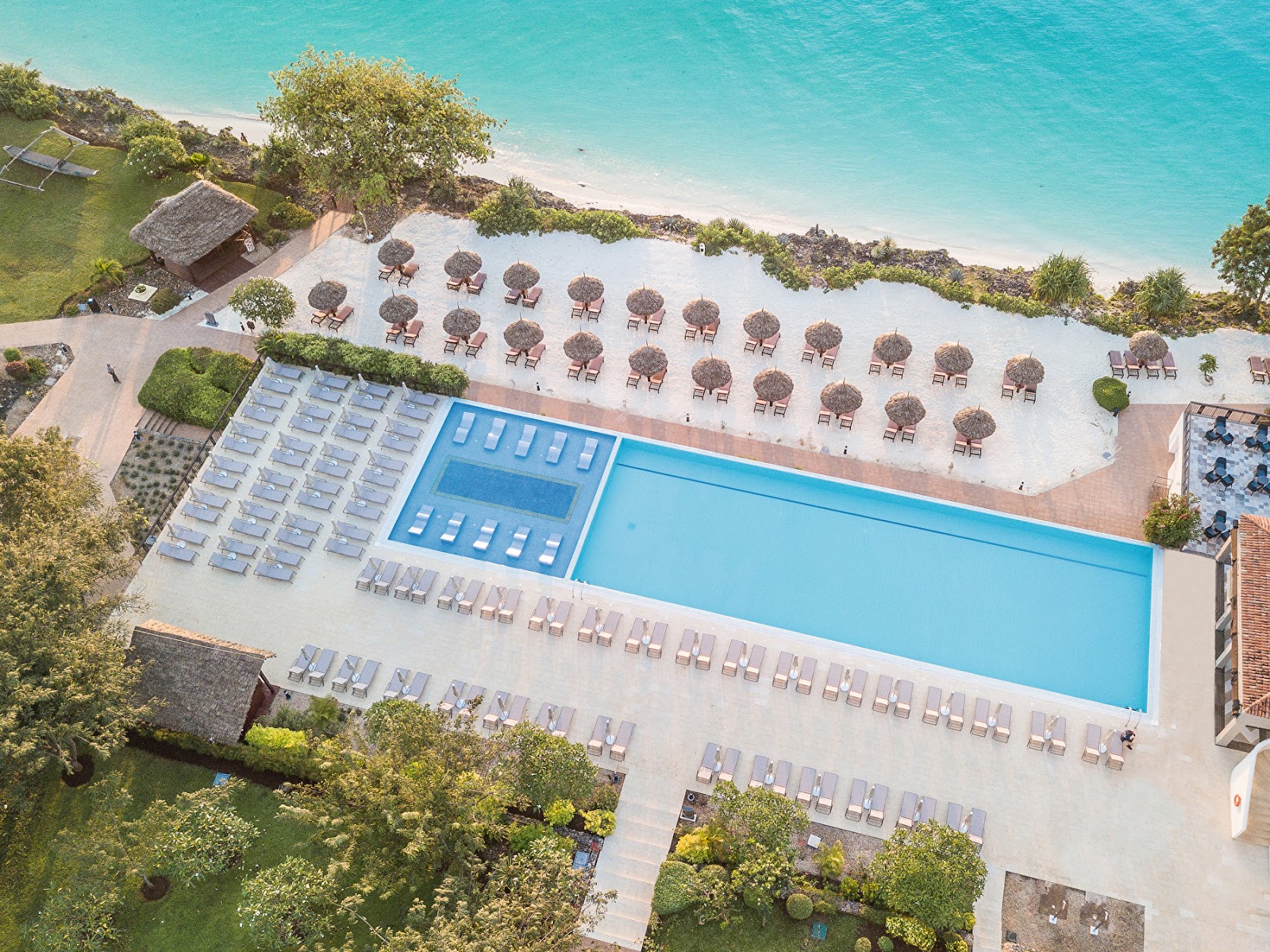 Aerial view of a resort pool and beach with rows of loungers and umbrellas overlooking turquoise water and white sand.