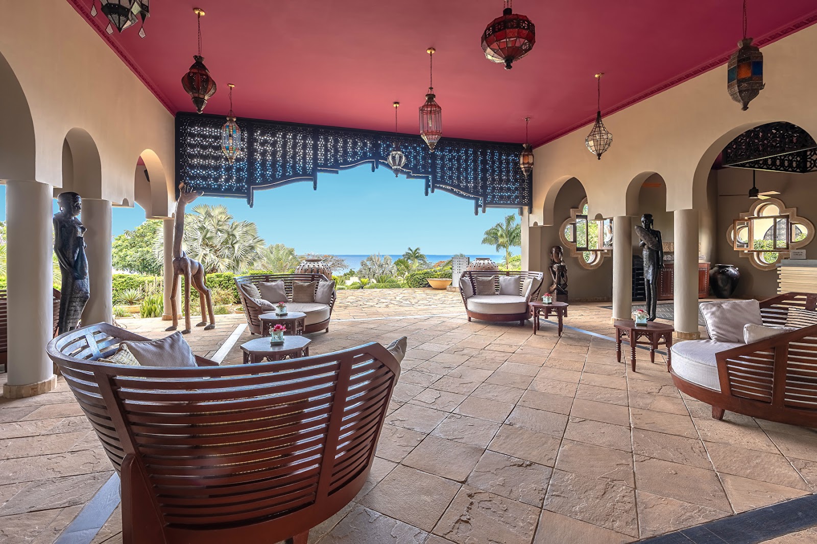 Covered terrace with pink ceiling, curved wooden seating, decorative lanterns, and ocean view beyond palm trees at Hotel Riu Palace Zanzibar.