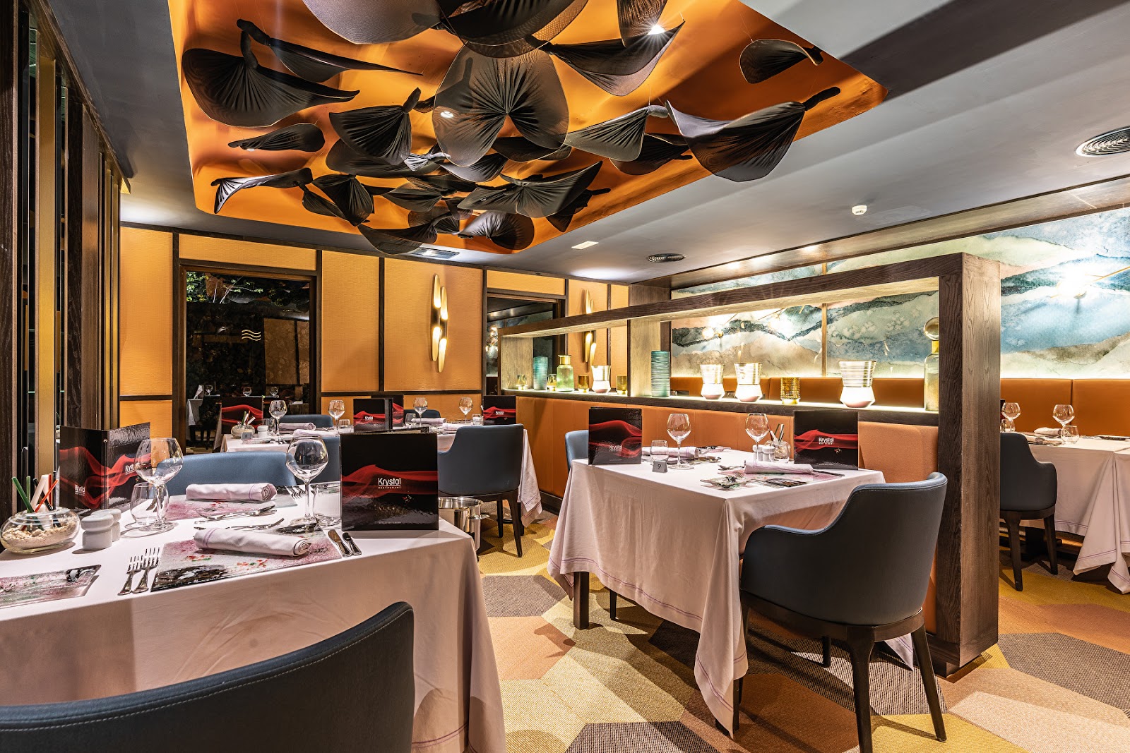 Fine dining restaurant with orange walls, white tablecloths, black chairs, and sculptural black leaf installations on the ceiling at Hotel Riu Palace Zanzibar.