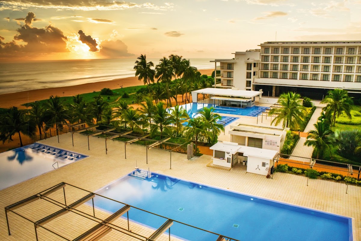 Resort pools and loungers overlook a sandy beach with palm trees and ocean at sunset.