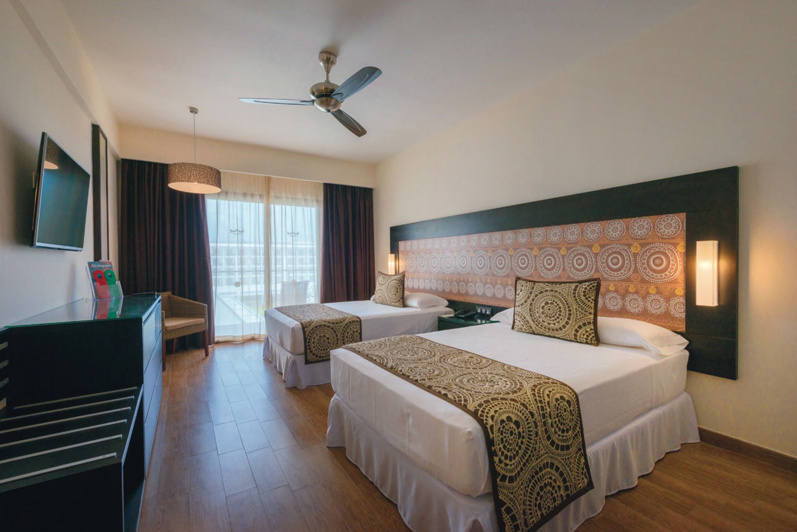 Resort room with two double beds, patterned headboards, dark curtains, ceiling fan, and balcony access at Hotel Riu Sri Lanka.