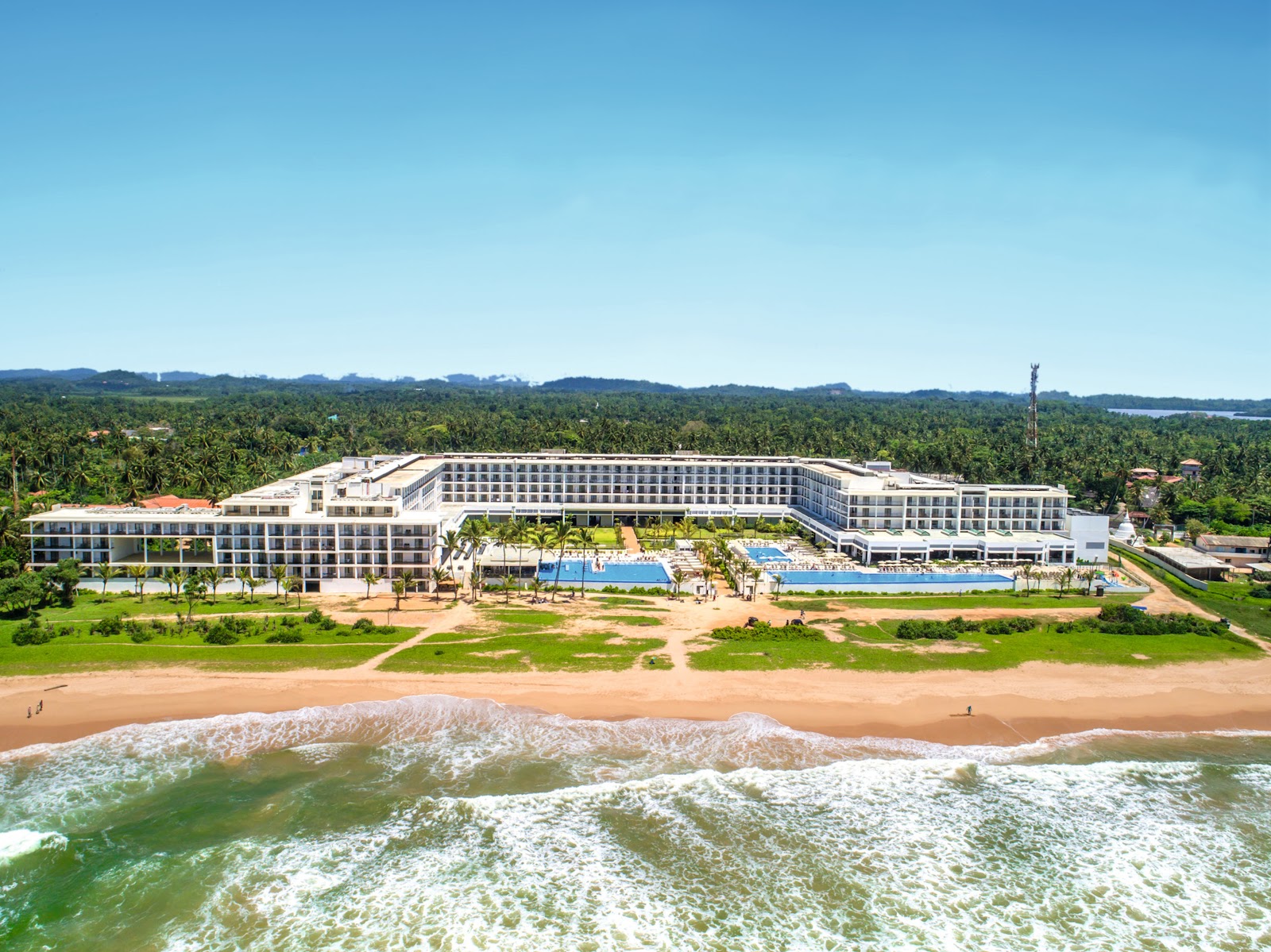 Poolside lounge chairs with umbrellas line the deck at Hotel Riu Sri Lanka, with the resort's blue-balconied buildings and a swimming pool visible beyond.