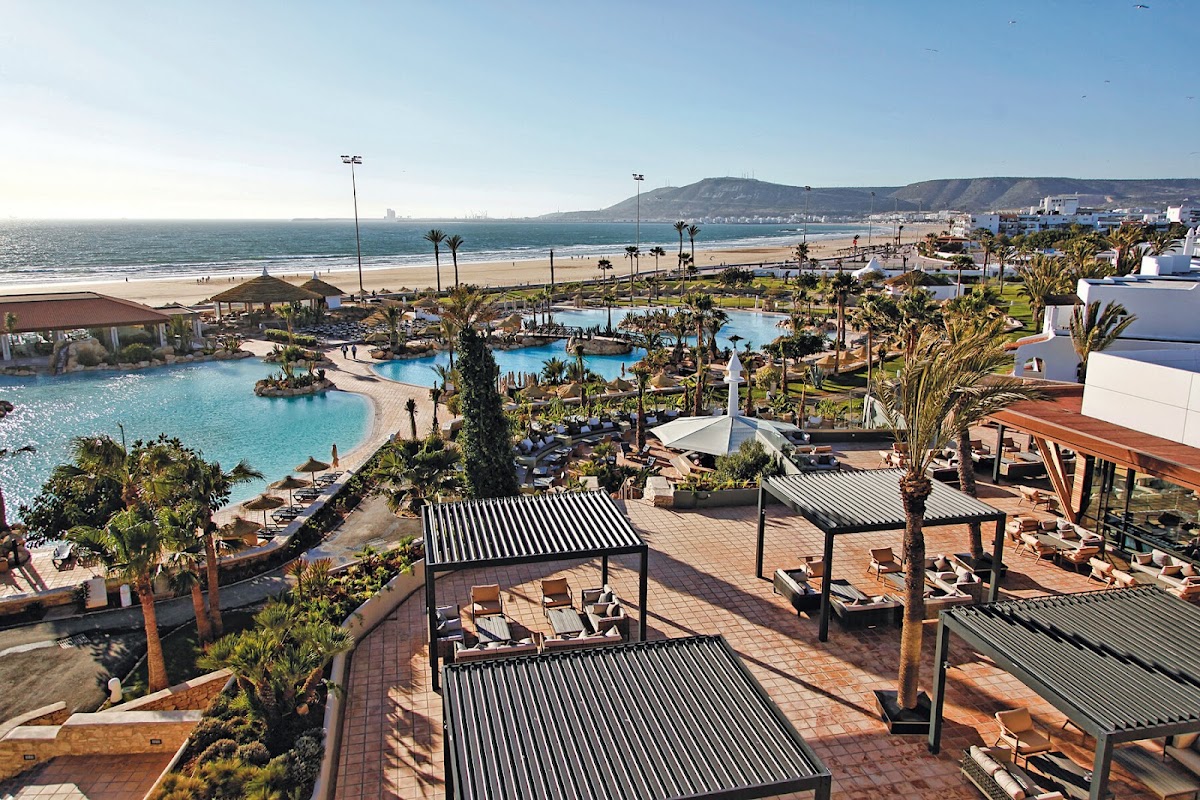 Terracotta terrace with striped shade structures overlooking lagoon pools, palm trees, and beach at Hotel Riu Tikida Dunas.