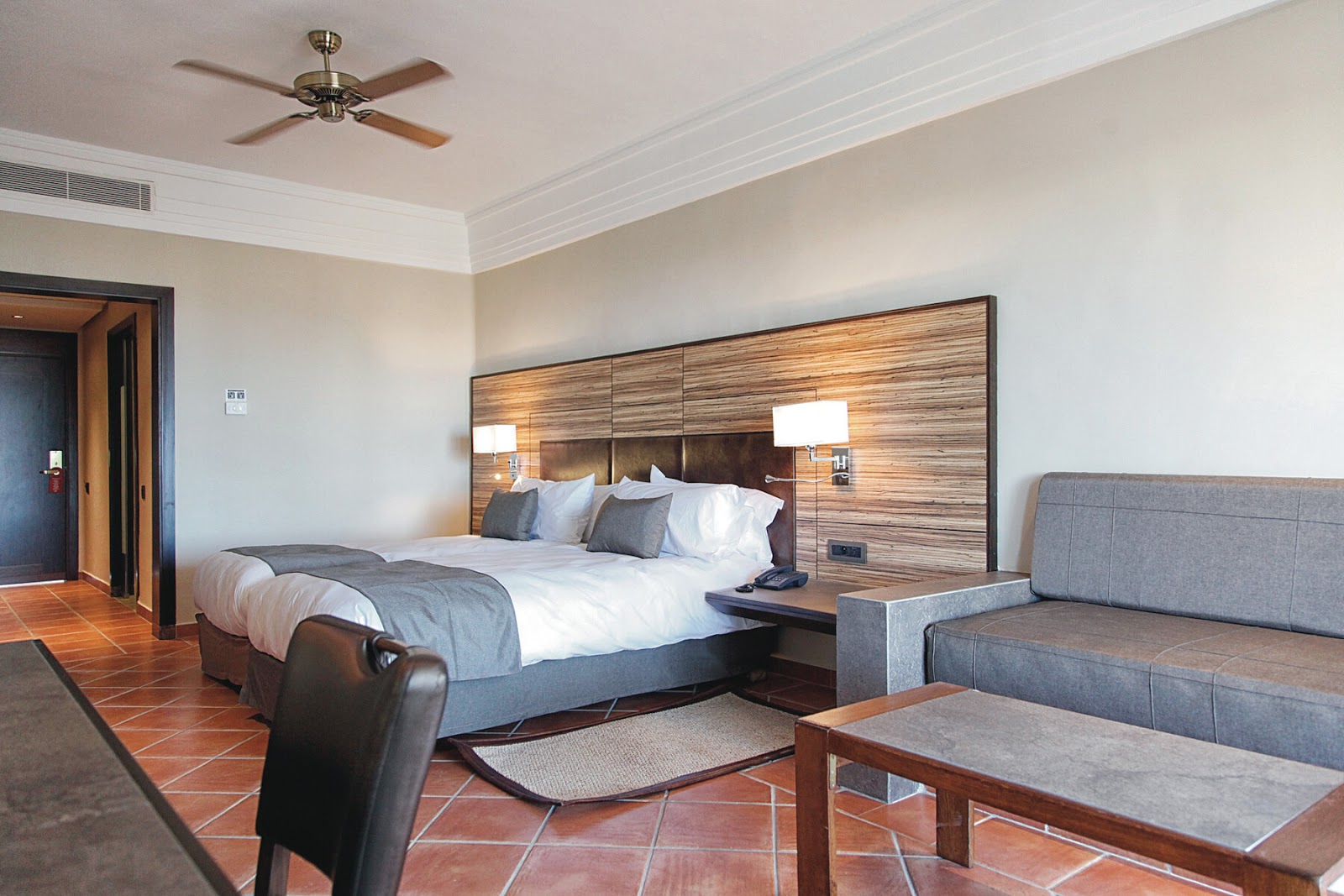 Resort bedroom with wood-paneled headboard, queen bed with white linens, gray sofa, ceiling fan, and terracotta tile flooring.