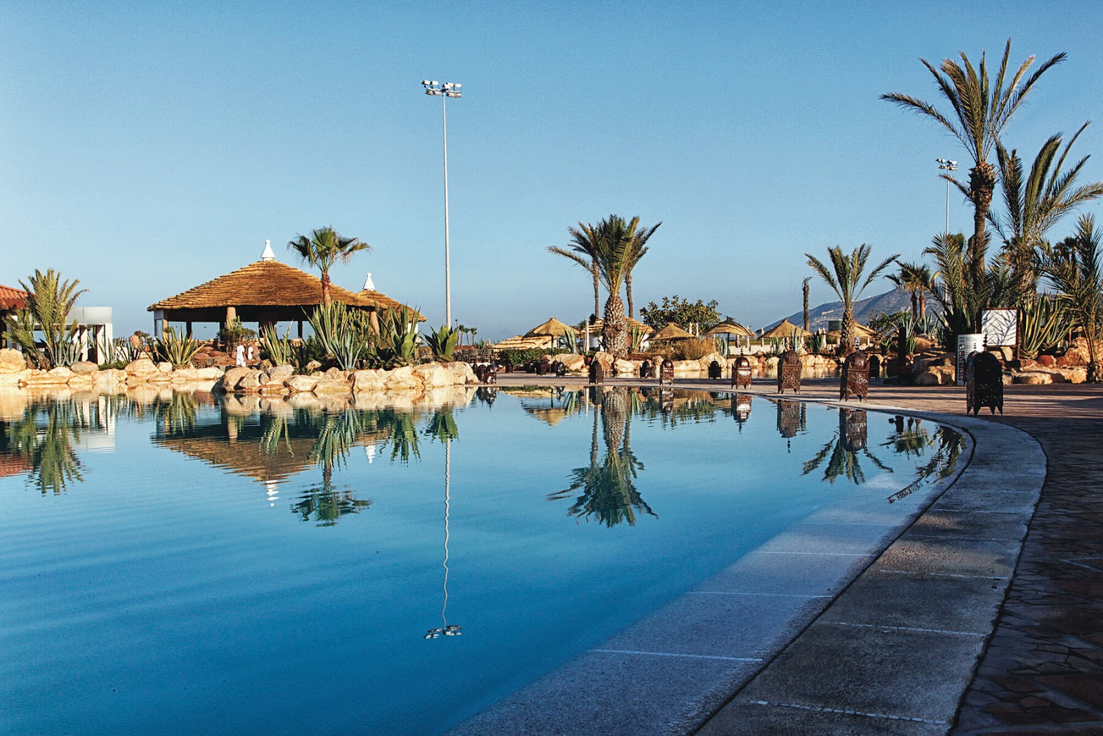 Pool with thatched-roof cabanas, palm trees, and guests at Hotel Riu Tikida Dunas under clear blue sky.