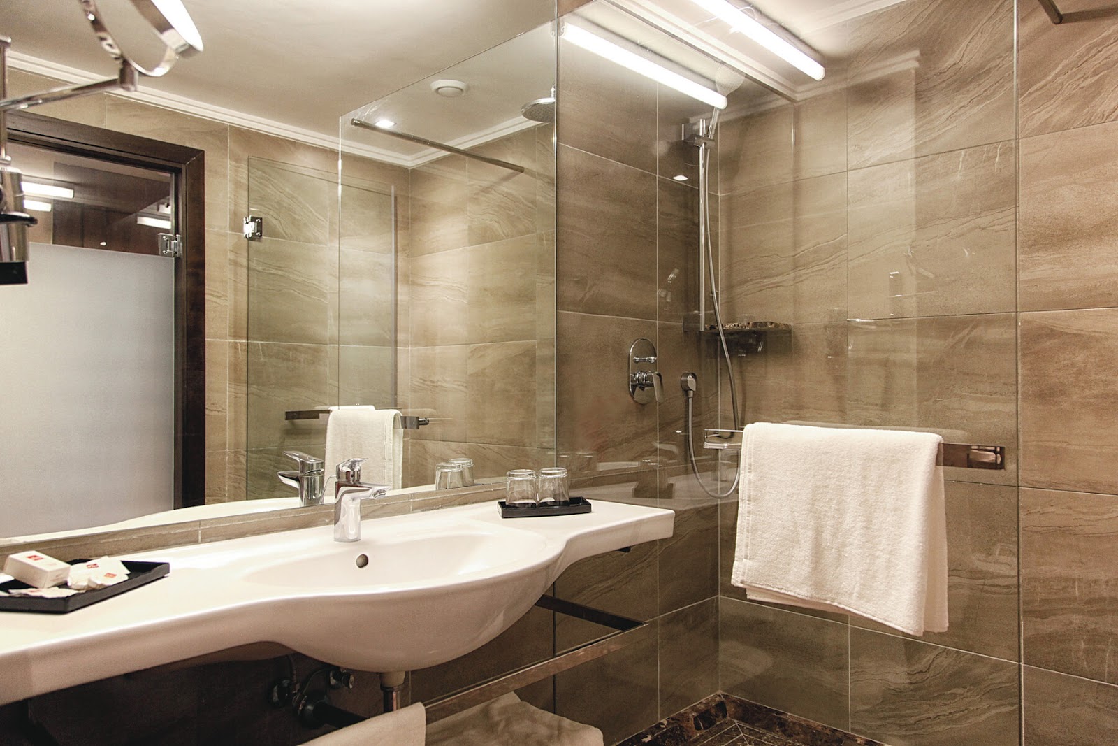Modern bathroom with curved white sink, large mirror, glass shower enclosure with beige tile walls, and white towels on racks.