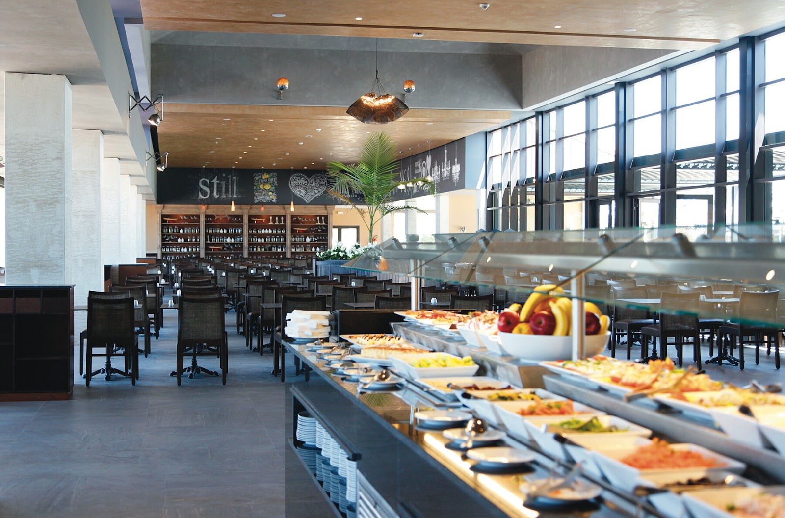 Modern dining room at Hotel Riu Tikida Dunas with buffet stations, black chairs, and floor-to-ceiling windows overlooking the landscape.