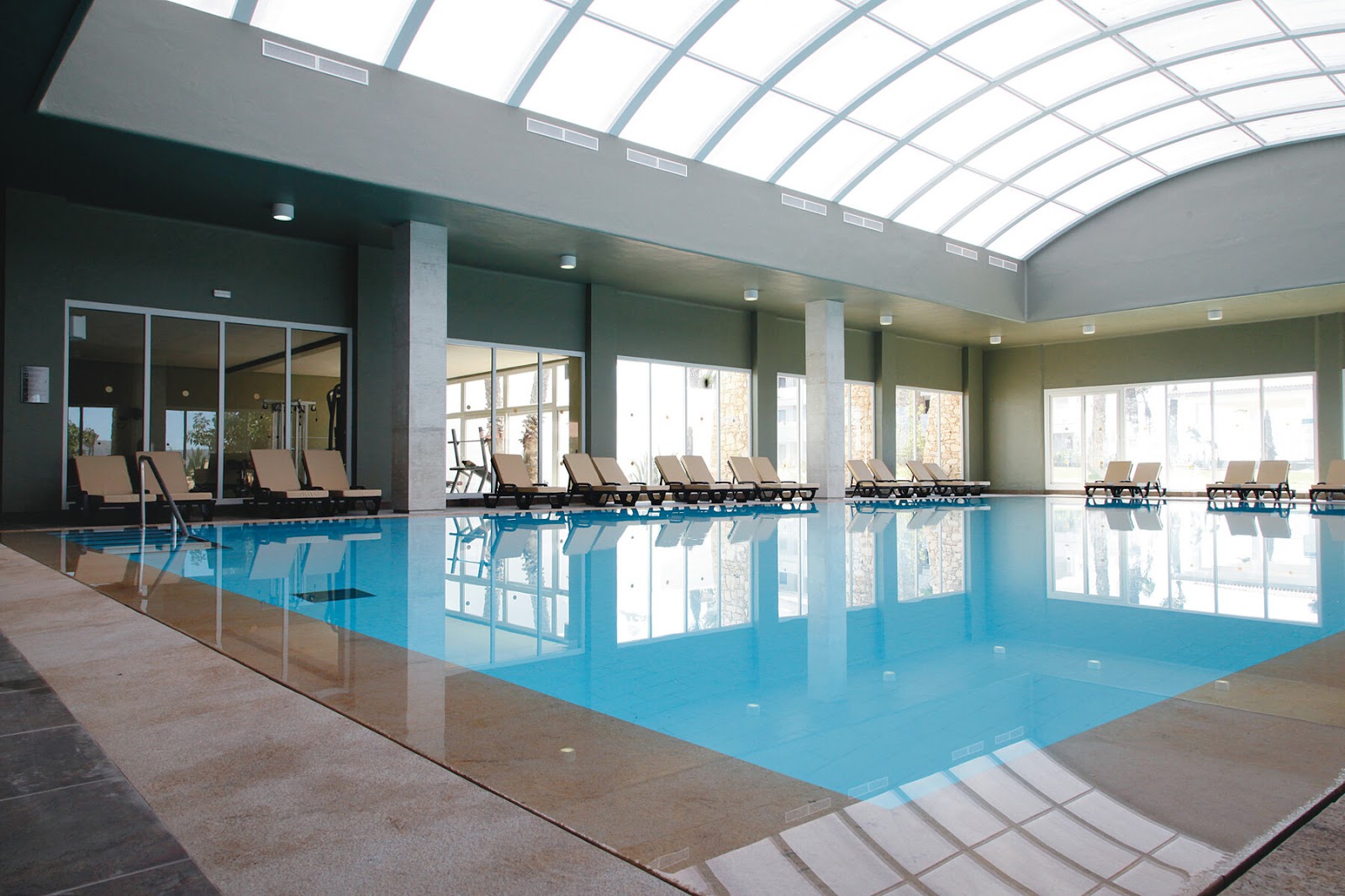 Indoor swimming pool at Hotel Riu Tikida Dunas with lounge chairs lining the sides and skylights illuminating the turquoise water.