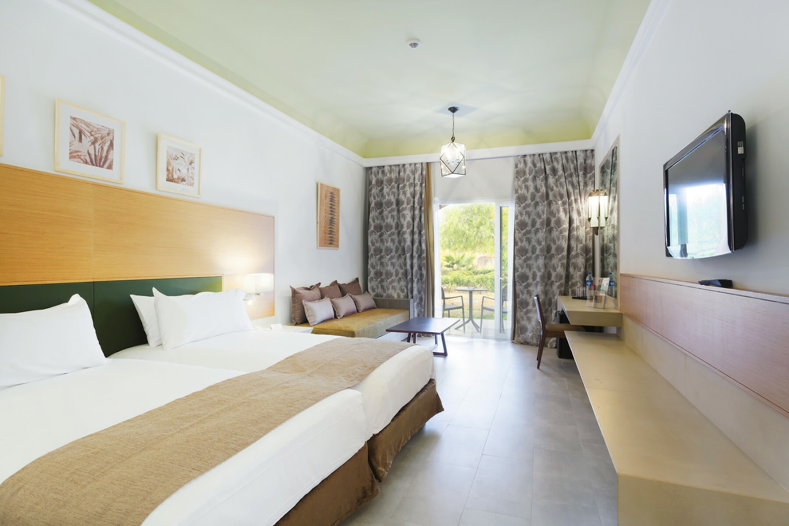 Resort bedroom with two beds, green headboards, patterned curtains, balcony access, TV, and pale green ceiling.
