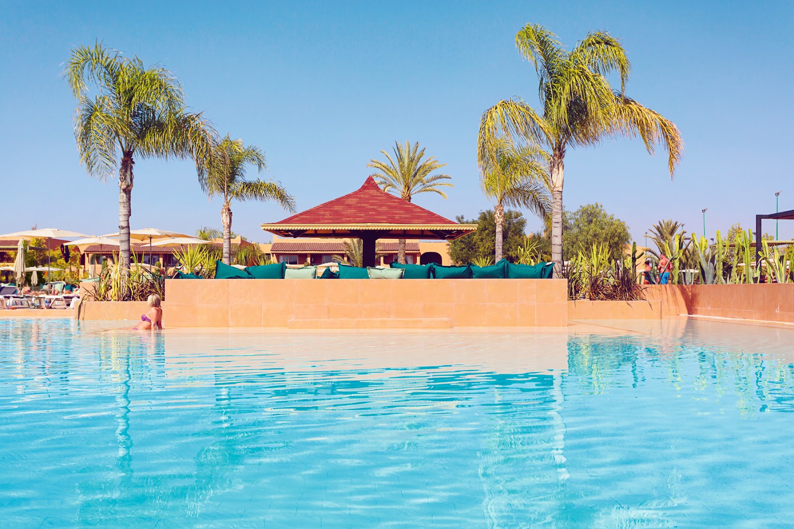 Turquoise pool with red-roofed pavilion, terracotta walls, and palm trees at Hotel Riu Tikida Palmeraie under clear sky.