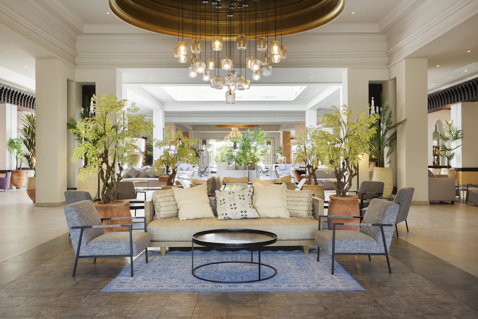 Spacious lobby with cream sofa, gray armchairs, potted plants, and a brass pendant chandelier overhead at Hotel Riu Tikida Palmeraie.