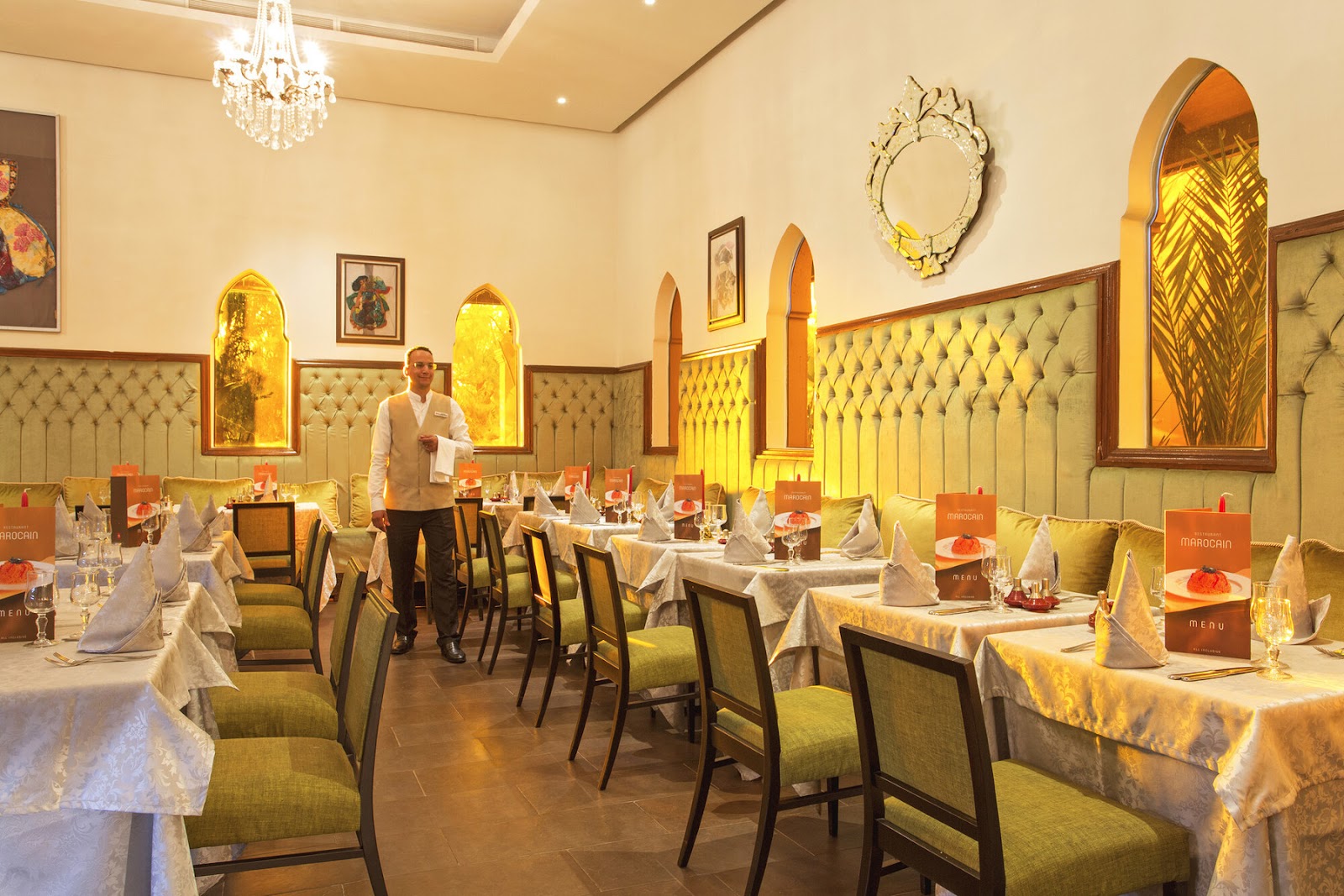 Upscale dining room with cream-colored tufted banquettes, white tablecloths, and gold-framed architectural details lining the walls under a crystal chandelier.