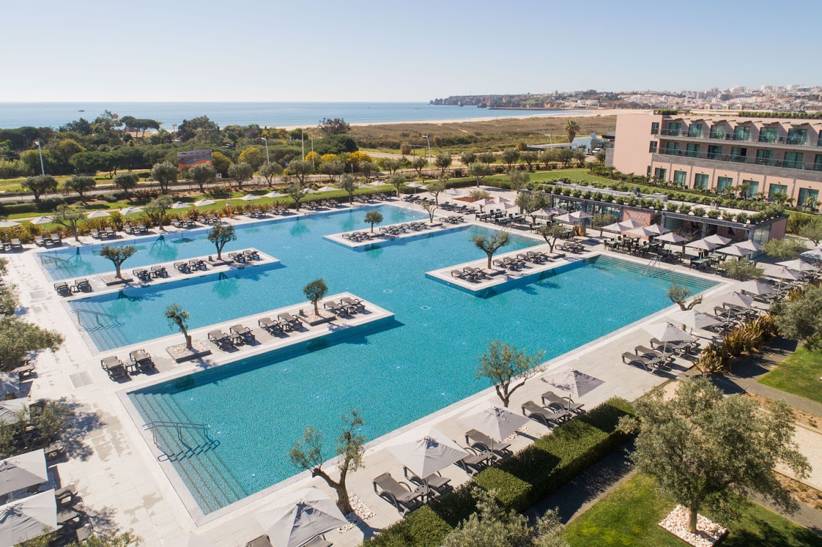 Multiple turquoise pools with loungers and islands of trees, coastal town visible beyond, pink hotel building on right overlooking landscaped grounds and beach.