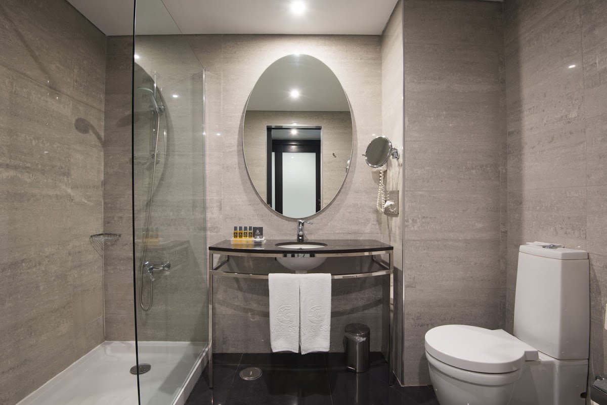 Modern bathroom with gray tile walls, oval mirror, dark vanity with white towels, glass shower enclosure, and toilet.