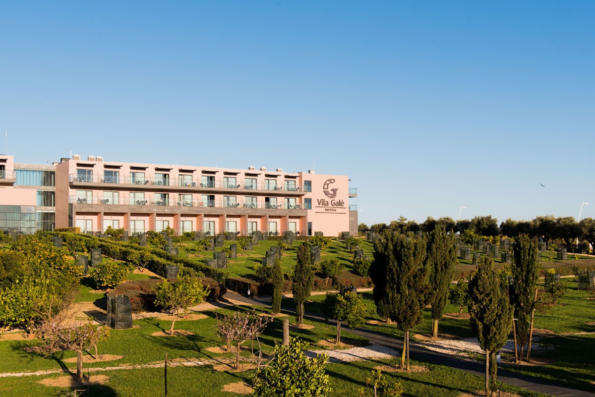Vila Galé Lagos hotel building with peachy-pink facade overlooking manicured grounds with cypress trees and garden pathways under clear blue sky.