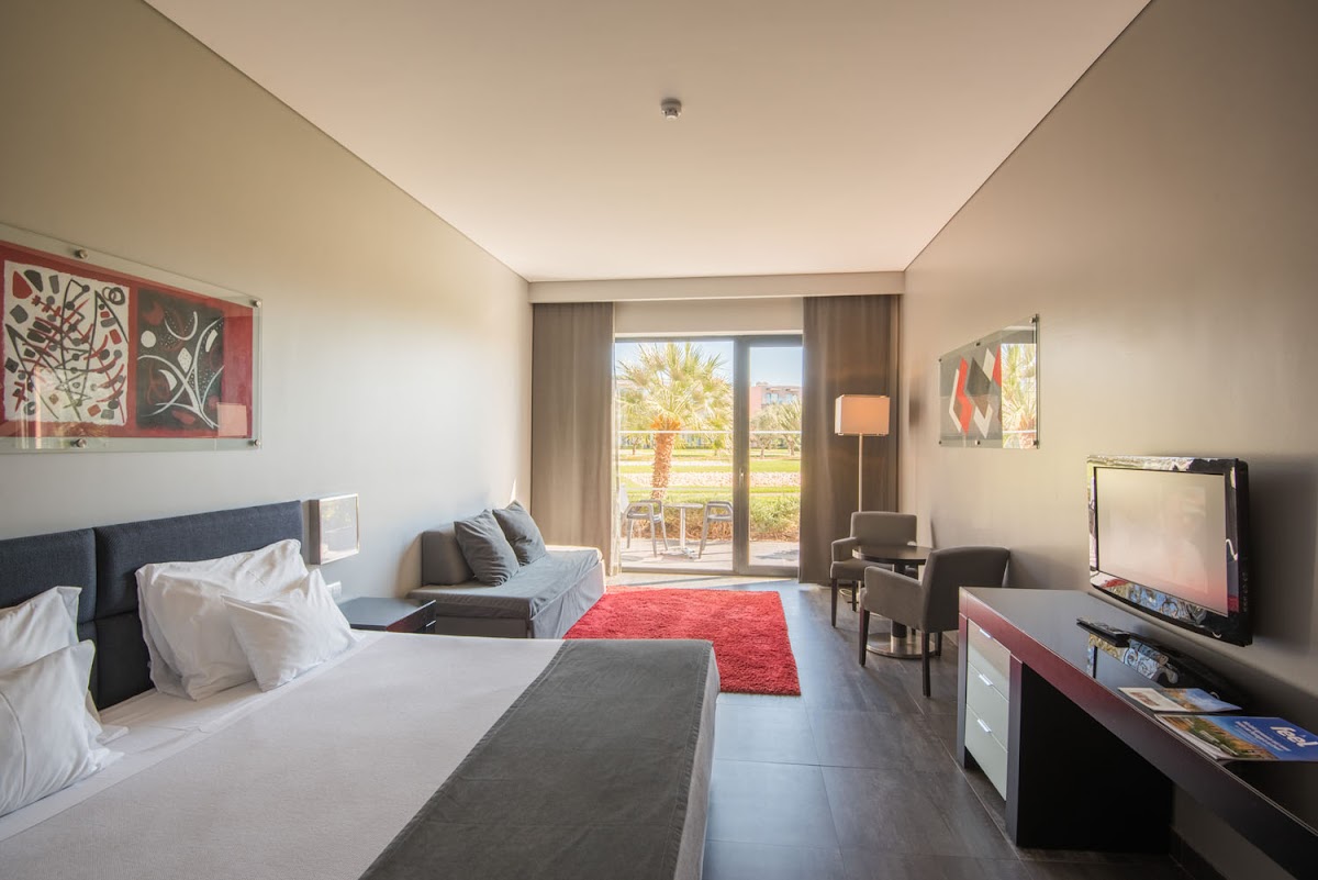 Spacious resort room with queen bed, sitting area, red accent rug, and glass doors opening to a balcony overlooking palm trees and grounds.