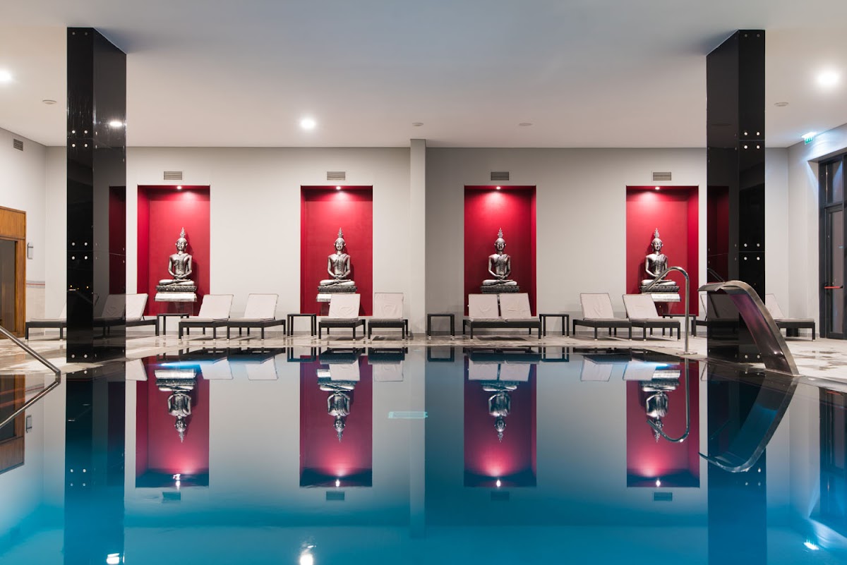 Indoor swimming pool with turquoise water, lounge chairs, and red-lit alcoves displaying Buddha statues at Hotel Vila Galé Lagos.