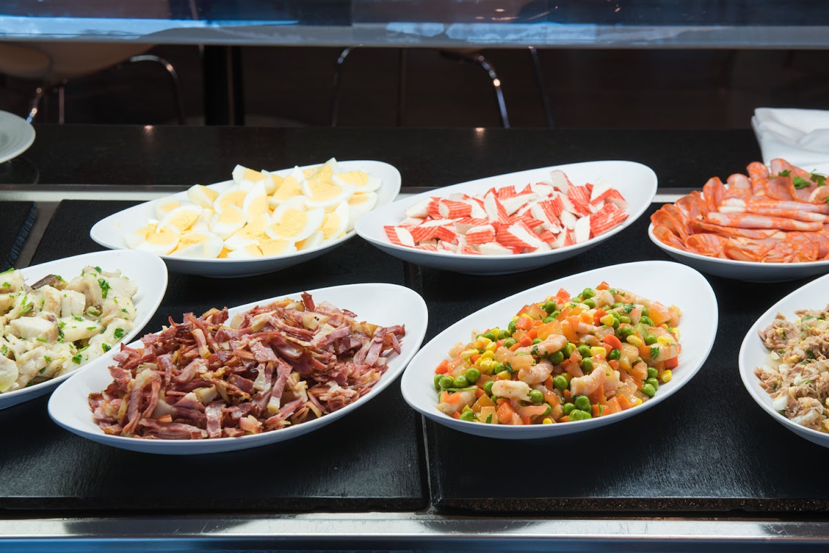 Buffet display at Hotel Vila Galé Lagos showing sliced cured meats, hard-boiled eggs, seafood, diced vegetables, and potato salad on white plates.