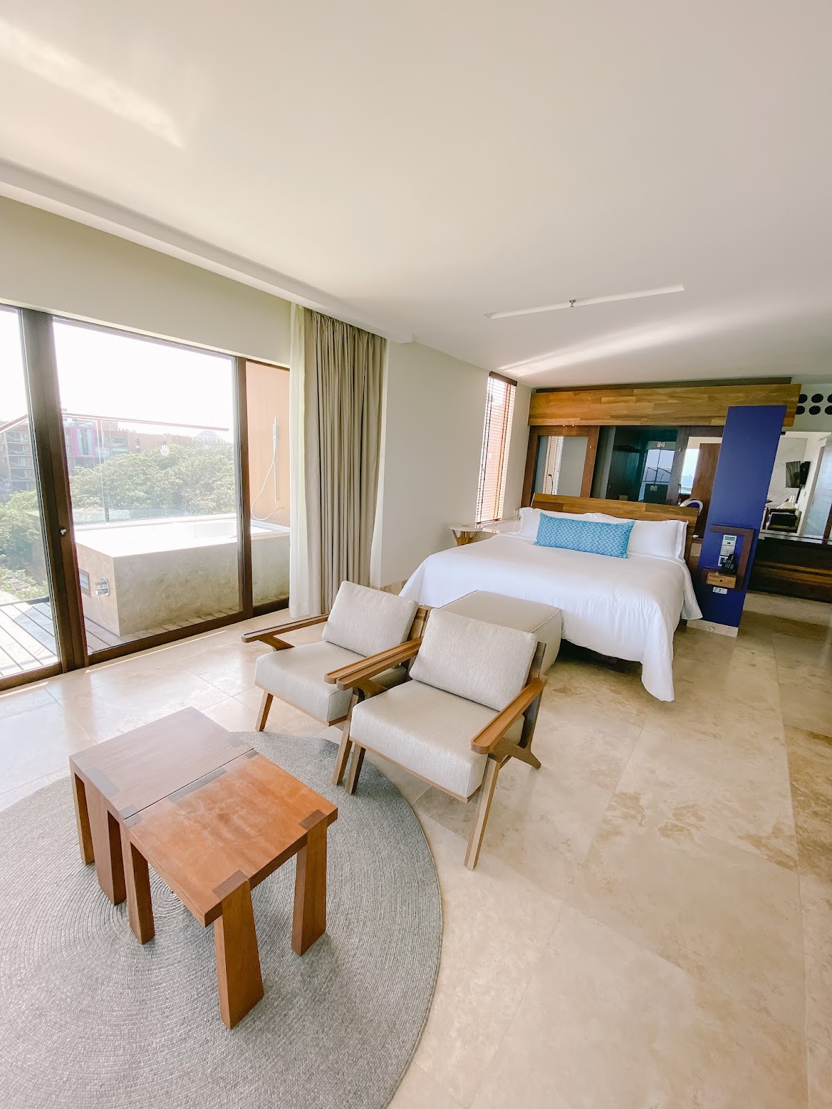 Modern resort suite with king bed, wooden lounge chairs, coffee table, and balcony overlooking landscape.