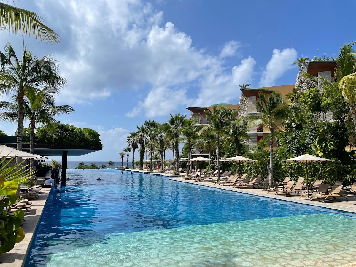 Long beachfront pool with pale umbrellas and loungers, flanked by palm trees and a multi-story stone building overlooking the ocean.
